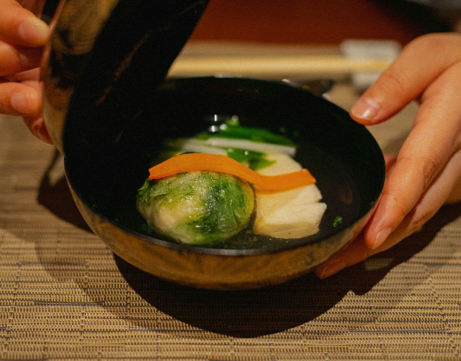 Two hands setting down a lacquer bowl of clear soup