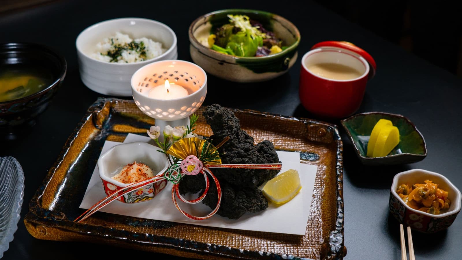 A candlelit black table set with small dishes, tea and a lacquered tray