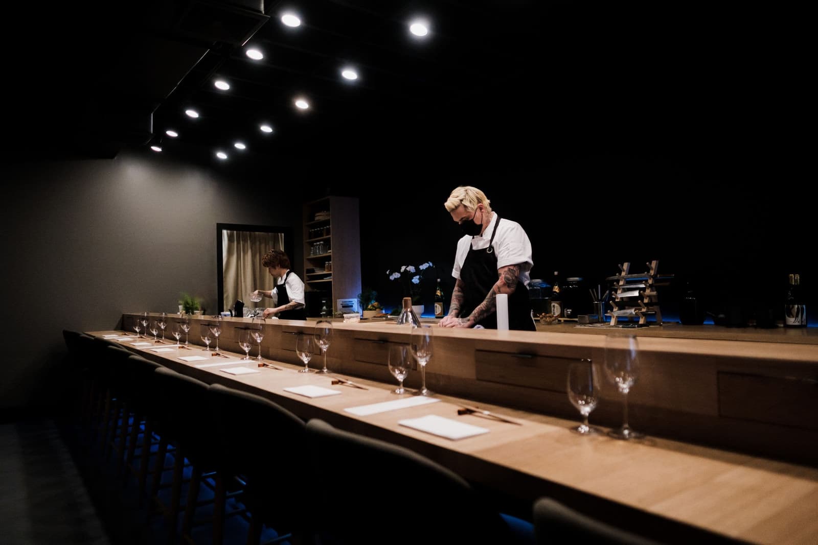 A long pale timber counter set with glasses under low spotlights, a chef at the far end